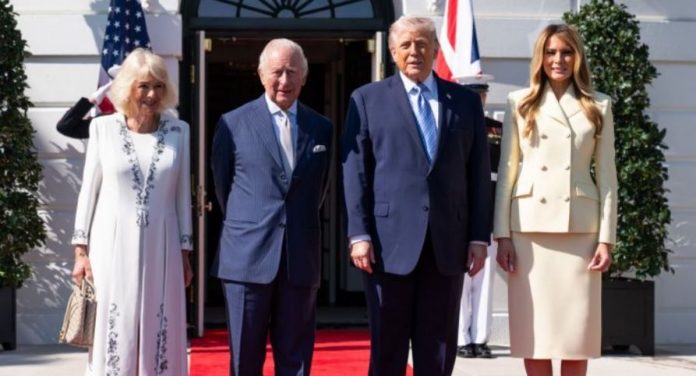 Two well-dressed couples stand on a red carpet outside a white building with flags, smiling for the camera.