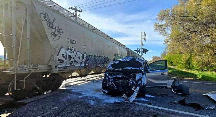 Car crash at a railroad crossing: black SUV with severe front-end damage beside a graffiti-covered freight train car, open door and scattered debris on the road
