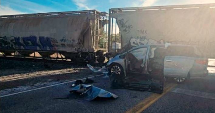 Silver pickup truck with major front-end damage and deployed airbags sits on a road beside two large freight railcars; a blue motorcycle is scattered in front of the truck, debris on the asphalt at a railroad crossing under a clear sky.