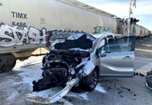 Silver sedan with severe front-end damage, hood up and door open, debris on the ground near graffiti-covered railcars at a train track.n