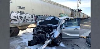 Silver sedan with severe front-end damage, hood up and door open, debris on the ground near graffiti-covered railcars at a train track.n