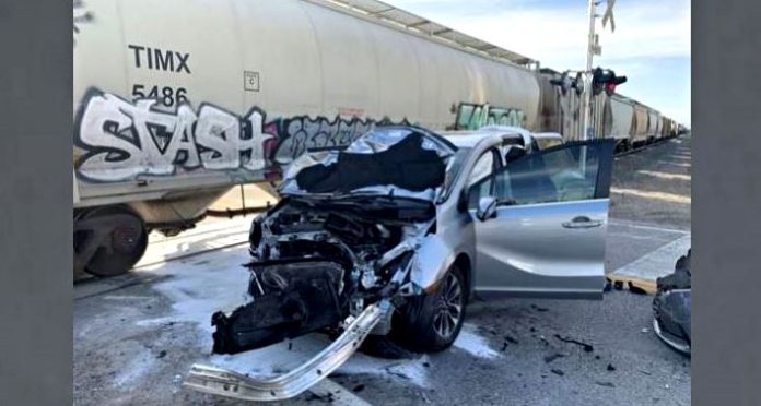 Silver sedan with severe front-end damage, hood up and door open, debris on the ground near graffiti-covered railcars at a train track.n