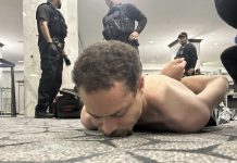 Shirtless man lying face down on carpeted floor with hands bound behind his back, surrounded by several police officers in tactical gear in a public indoor space.