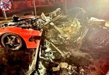 Severely damaged, burned red sports car wreck with burnt debris and charred rubble scattered on the ground; emergency responders and a fire truck are visible in the background at night.