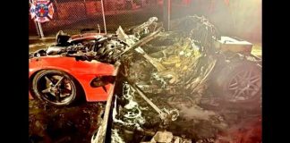 Severely damaged, burned red sports car wreck with burnt debris and charred rubble scattered on the ground; emergency responders and a fire truck are visible in the background at night.