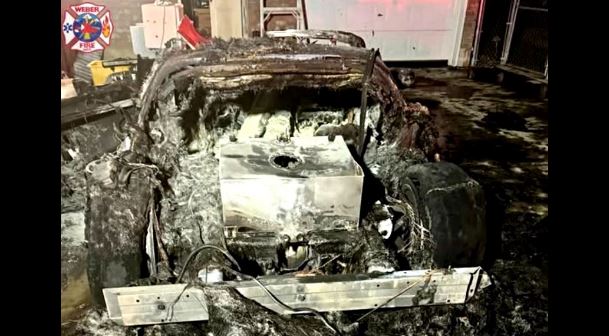 Charred car exposed to a severe fire, with a melted interior and scorched exterior in a workshop setting, emergency logo visible in the corner.