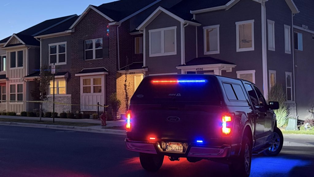 Emergency vehicle with red and blue lights parked on a residential street at night, yellow caution tape visible by the house.