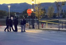 Group of police officers standing in a street at dusk near a Stop sign and crime scene tape. A vehicle is parked on the left.