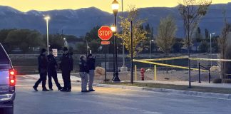 Group of police officers standing in a street at dusk near a Stop sign and crime scene tape. A vehicle is parked on the left.
