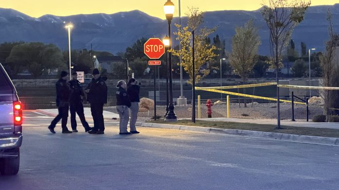 Group of police officers standing in a street at dusk near a Stop sign and crime scene tape. A vehicle is parked on the left.