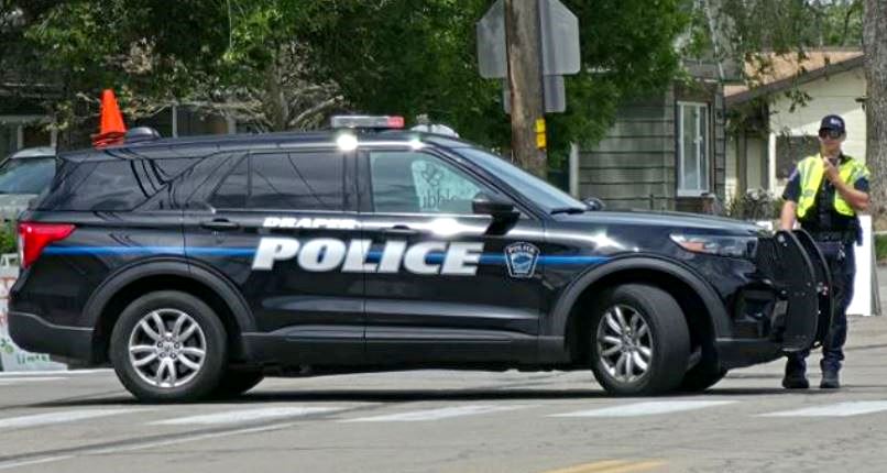 Dark police SUV with'POLICE' lettering blocks a street; an officer in a reflective vest stands nearby.