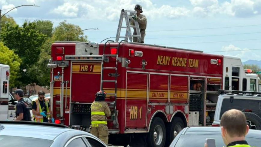 Fire department heavy rescue truck with firefighters around and a worker on a ladder atop the vehicle, outdoors near cars and trees