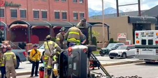 Emergency responders and firefighters work around a flipped SUV on a city street; an ambulance is nearby as bystanders watch.