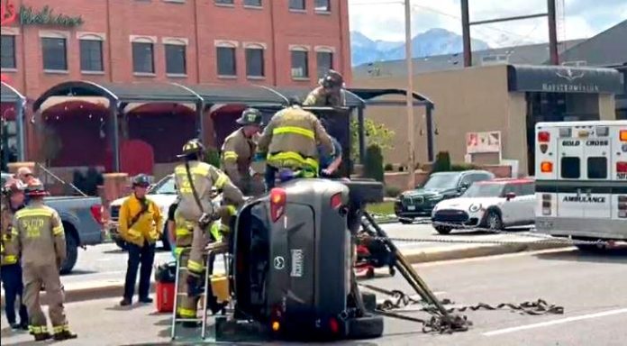 Emergency responders and firefighters work around a flipped SUV on a city street; an ambulance is nearby as bystanders watch.
