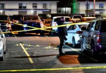 Nighttime parking lot cordoned off with yellow police tape; a person stands by a white car as evidence markers dot the pavement.