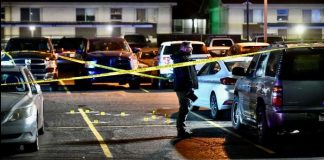 Nighttime parking lot cordoned off with yellow police tape; a person stands by a white car as evidence markers dot the pavement.