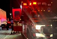 Emergency responders with a red-flashing ambulance on a dark street; a person stands near a white truck beside the vehicle doors open.