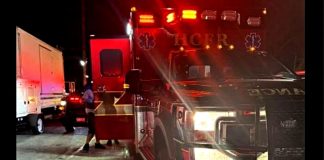 Emergency responders with a red-flashing ambulance on a dark street; a person stands near a white truck beside the vehicle doors open.