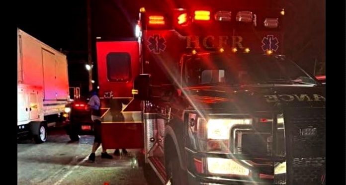 Emergency responders with a red-flashing ambulance on a dark street; a person stands near a white truck beside the vehicle doors open.