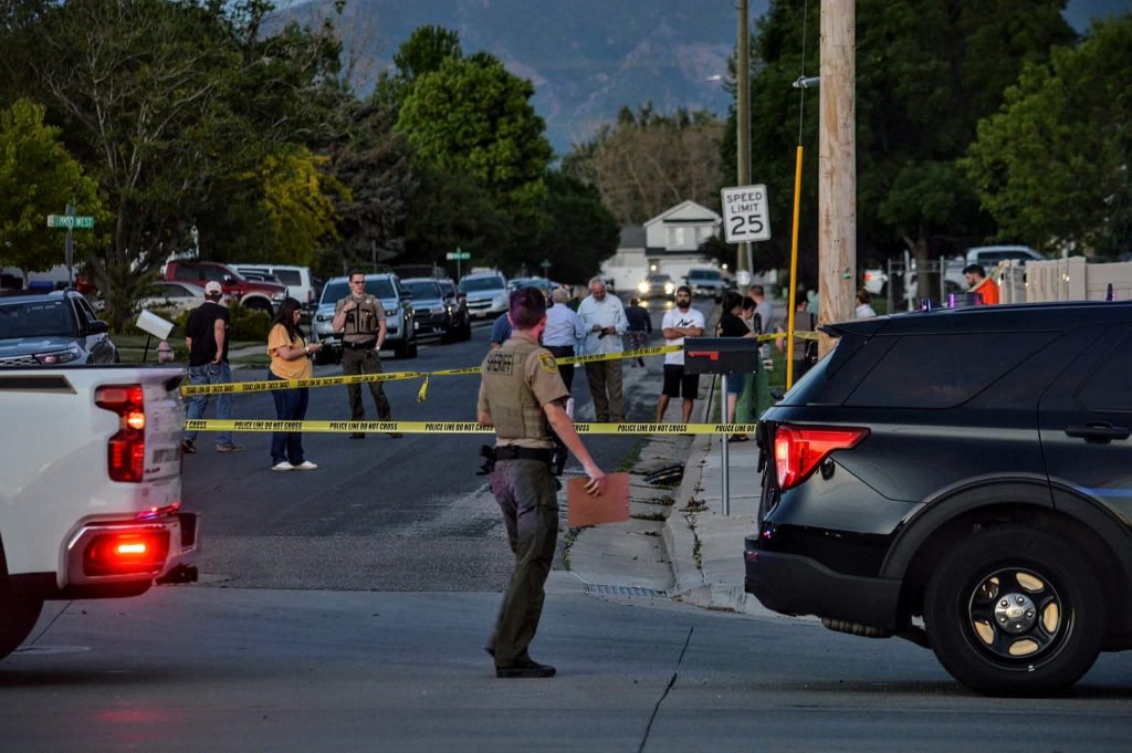 Street blocked by police tape and emergency responders on a residential road; several officers and parked cars in the background.