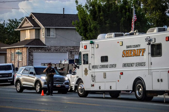Davis County Sheriff mobile command center vehicle parked on a city street beside a cone and several cars, with a two-story house in the background and people nearby.