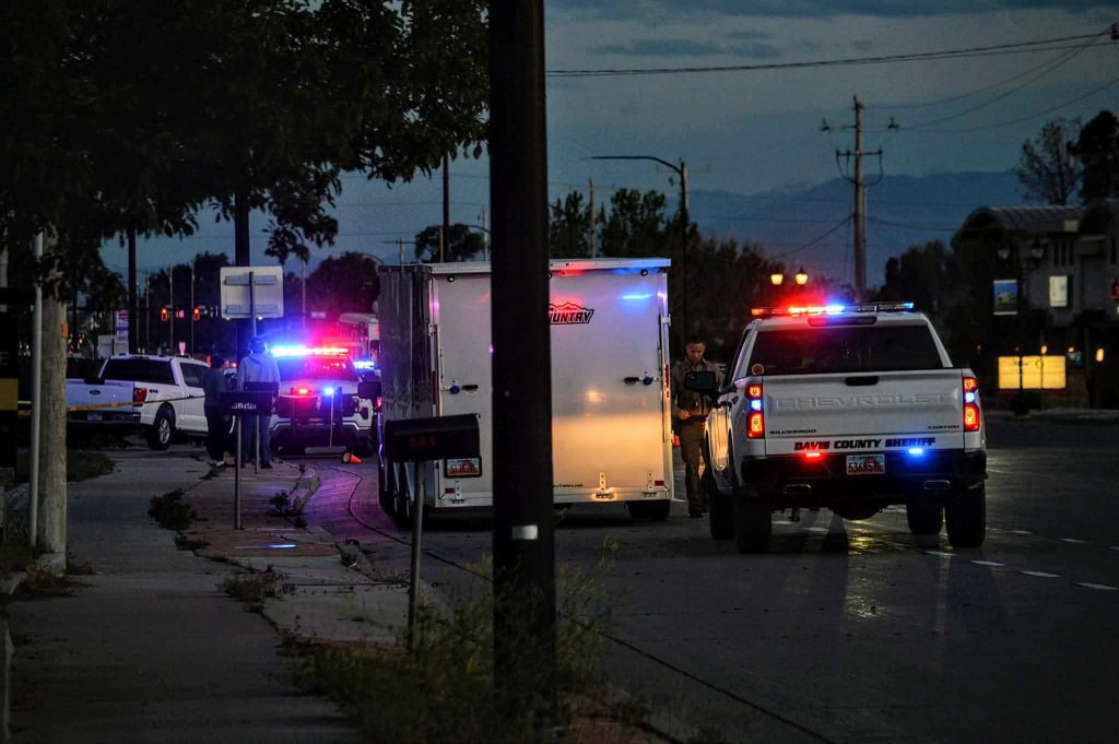 Emergency scene at dusk with multiple police vehicles and flashing red/blue lights on a street, a white utility truck in the foreground, and deputies assisting nearby.