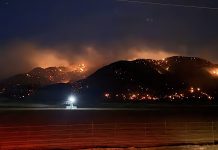 Night scene of a wildfire burning along a hill ridge, with flames and smoke across the slope and a bright light on the foreground road barrier.