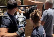 People outdoors around a metal railing; a man with a cordless angle grinder operates the tool while others watch, wearing gloves and navy shirts.