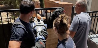 People outdoors around a metal railing; a man with a cordless angle grinder operates the tool while others watch, wearing gloves and navy shirts.