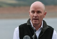 Bald man speaking at a microphone outdoors, wearing a dark vest with an emblem on it.