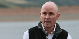 Bald man speaking at a microphone outdoors, wearing a dark vest with an emblem on it.