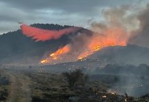 Helicopter flies over a hillside wildfire with orange flames and dark smoke across the ridge.