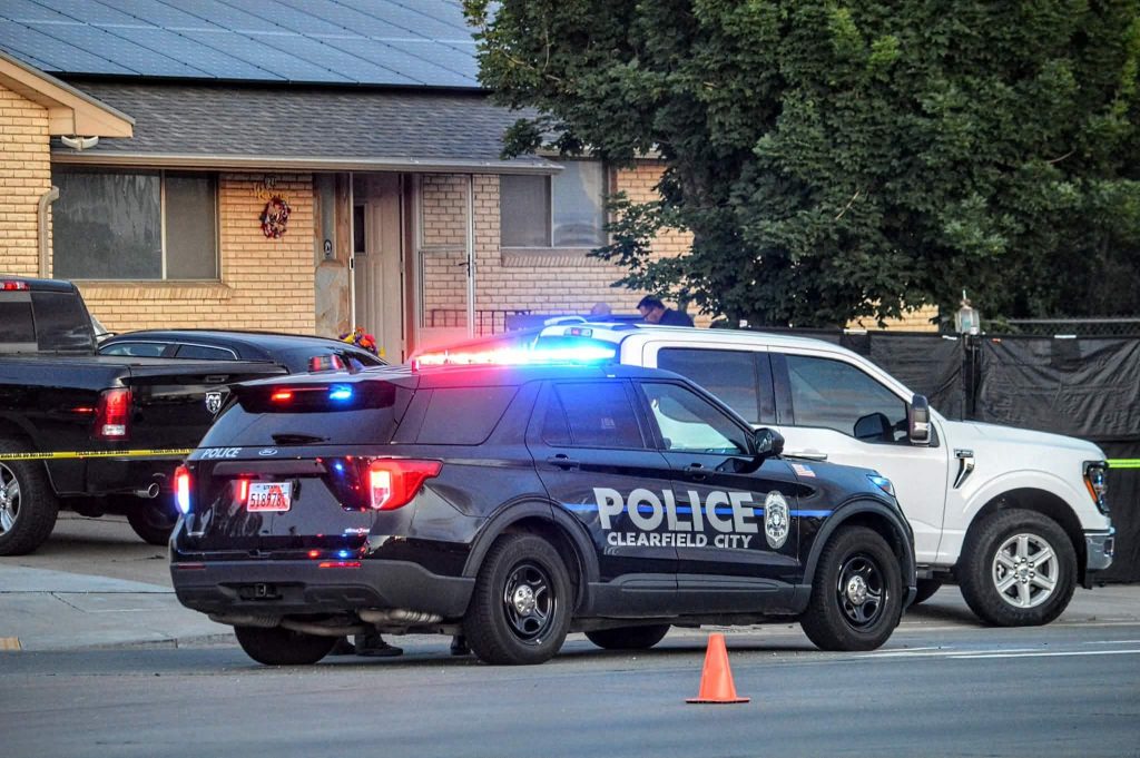 Police cars with flashing red/blue lights parked in front of a brick house; orange cone and caution tape on the street nearby.