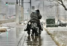 Person in a wheelchair being pushed by a companion on a wet sidewalk as it snows in an urban street.