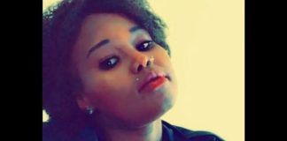 Portrait of a young woman with short curly hair, nose piercing, red lipstick, tilting her head slightly to the side.
