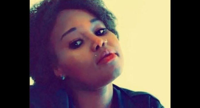 Portrait of a young woman with short curly hair, nose piercing, red lipstick, tilting her head slightly to the side.