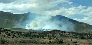 Smoke plumes rise from a brush fire in a dry grassland with mountains in the background under a blue sky.