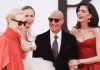 Four adults posing together on a red carpet at a formal event: two women in red gowns, one in a champagne gown, and a bald man in a suit with sunglasses.