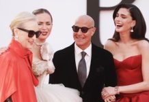 Four adults posing together on a red carpet at a formal event: two women in red gowns, one in a champagne gown, and a bald man in a suit with sunglasses.