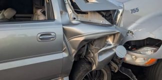 Silver pickup truck with heavy front-end damage from a collision with a large gray truck, driver-side area crumpled and wheel exposed