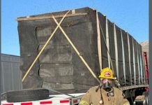 Firefighter in turnout gear with respirator stands near a large, black-wrapped cargo crate strapped to a flatbed trailer.