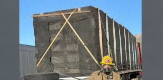 Firefighter in turnout gear with respirator stands near a large, black-wrapped cargo crate strapped to a flatbed trailer.