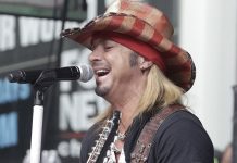 Male guitarist singing into a microphone on stage, wearing a red plaid cowboy hat and black shirt.