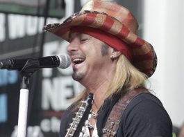 Male guitarist singing into a microphone on stage, wearing a red plaid cowboy hat and black shirt.