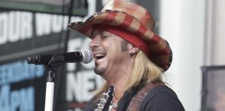 Male guitarist singing into a microphone on stage, wearing a red plaid cowboy hat and black shirt.