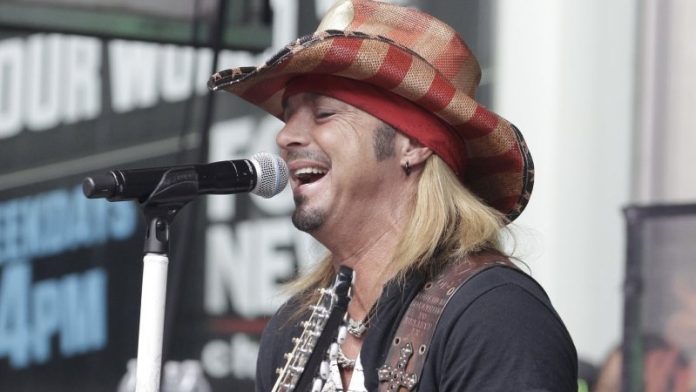 Male guitarist singing into a microphone on stage, wearing a red plaid cowboy hat and black shirt.
