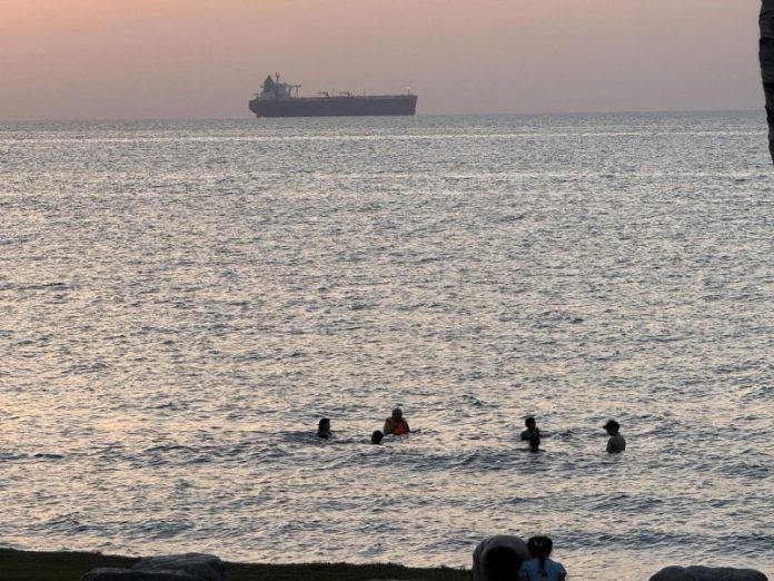 Britain-pledges-drones-jets-warship-to-Strait-of-Hormuz-mission-1 Five swimmers in the sea at sunset near a rocky shore, with a large cargo ship on the horizon.