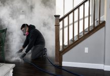 Worker in safety gear kneels on a dark wood floor, operating a fogging machine that releases dense vapor through a blue hose in a living room near a staircase.