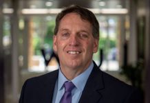 Smiling middle-aged man in a navy suit and purple tie standing in a modern office lobby.