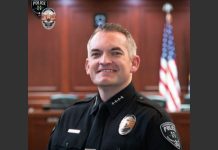 Police officer in uniform smiling in a courtroom with an American flag in the background and badge visible on chest.
